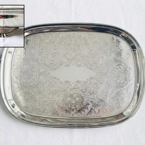 Pu&ograve; includere: Un vassoio da portata ovale color argento con intricati motivi floreali e a volute in rilievo sulla superficie, un'area centrale liscia e due manici decorati sui lati. L'inserto mostra un primo piano dei piedini metallici curvi.