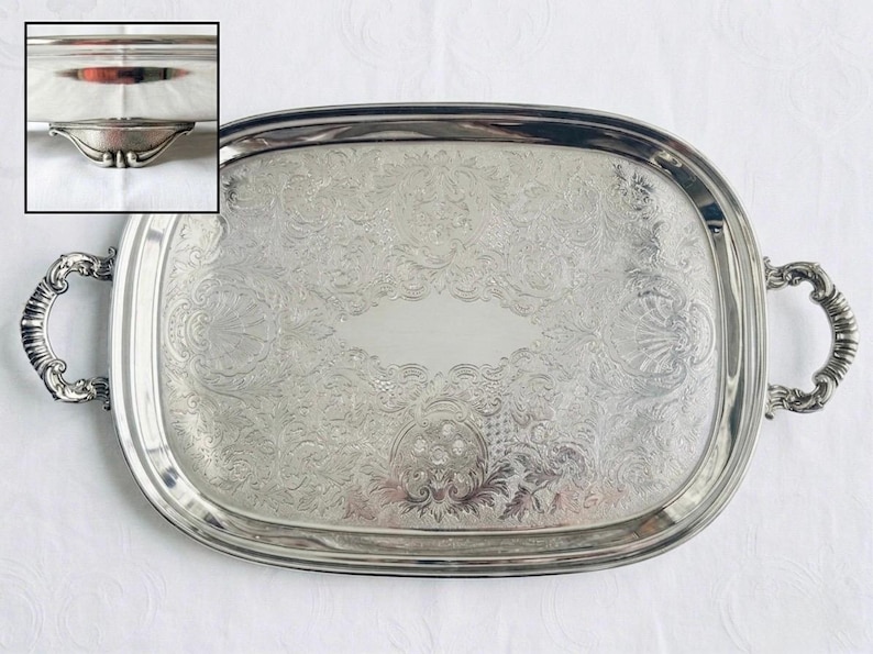Pu&ograve; includere: Un vassoio da portata ovale color argento con intricati motivi floreali e a volute in rilievo sulla superficie, un'area centrale liscia e due manici decorati sui lati. L'inserto mostra un primo piano dei piedini metallici curvi.