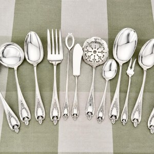 May include: A collection of eleven silver-toned metal utensils featuring ornate floral detailing on the handles. The set includes various spoons, forks, a butter knife, a pierced serving spoon, and a small two-pronged fork arranged in a row.