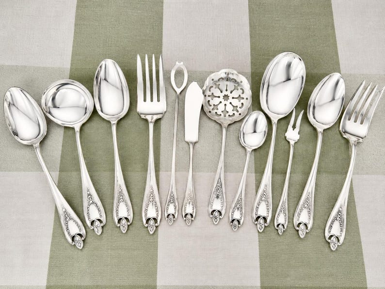 May include: A collection of eleven silver-toned metal utensils featuring ornate floral detailing on the handles. The set includes various spoons, forks, a butter knife, a pierced serving spoon, and a small two-pronged fork arranged in a row.