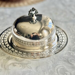May include: A silver-colored butter dish with a domed lid and ornate handle. The dish sits on a matching, intricately patterned plate. The reflective surface of the lid mirrors the surroundings, creating a vintage aesthetic.