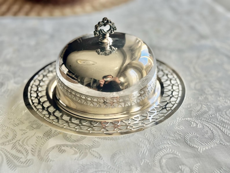 May include: A silver-colored butter dish with a domed lid and ornate handle. The dish sits on a matching, intricately patterned plate. The reflective surface of the lid mirrors the surroundings, creating a vintage aesthetic.