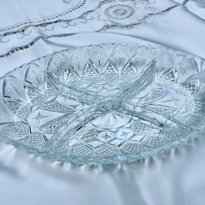 May include: Clear, oval-shaped glass serving dish with three divided sections. The dish features a detailed, textured pattern of diamond shapes and starbursts. The dish is on a white tablecloth.