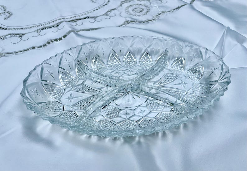May include: Clear, oval-shaped glass serving dish with three divided sections. The dish features a detailed, textured pattern of diamond shapes and starbursts. The dish is on a white tablecloth.