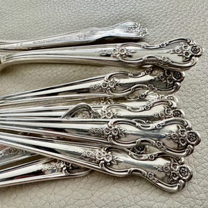 May include: Close-up of a collection of ornate silver spoons. The spoons have a floral design on the handles and are arranged in a layered pattern. The spoons are made of a shiny, reflective metal.