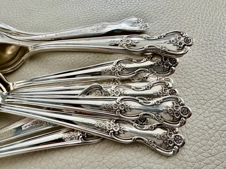 May include: Close-up of a collection of ornate silver spoons. The spoons have a floral design on the handles and are arranged in a layered pattern. The spoons are made of a shiny, reflective metal.