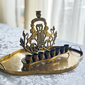 May include: A gold-toned menorah with a decorative lion and Torah design, set on an oval tray. The menorah holds eight small cups for candles. The tray has handles and a textured surface.