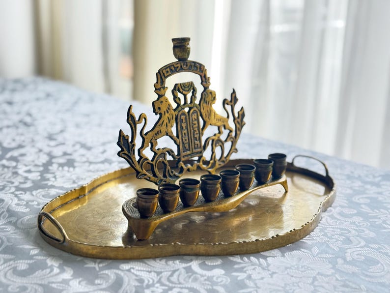 May include: A gold-toned menorah with a decorative lion and Torah design, set on an oval tray. The menorah holds eight small cups for candles. The tray has handles and a textured surface.