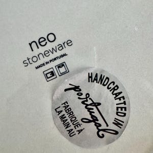 May include: A light-coloured stoneware surface featuring the black printed text neo stoneware MADE IN PORTUGAL with two square icons. A circular sticker is applied, reading HANDCRAFTED IN Portugal and FABRIQU&Eacute; &Agrave; LA MAIN AU in black text.