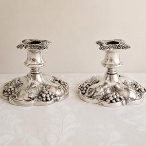 May include: A pair of silver-toned metal candle holders featuring a wide, scalloped base decorated with embossed grape clusters and vine leaf patterns. Each holder has a central stem and a square-shaped top rim with detailed floral accents.