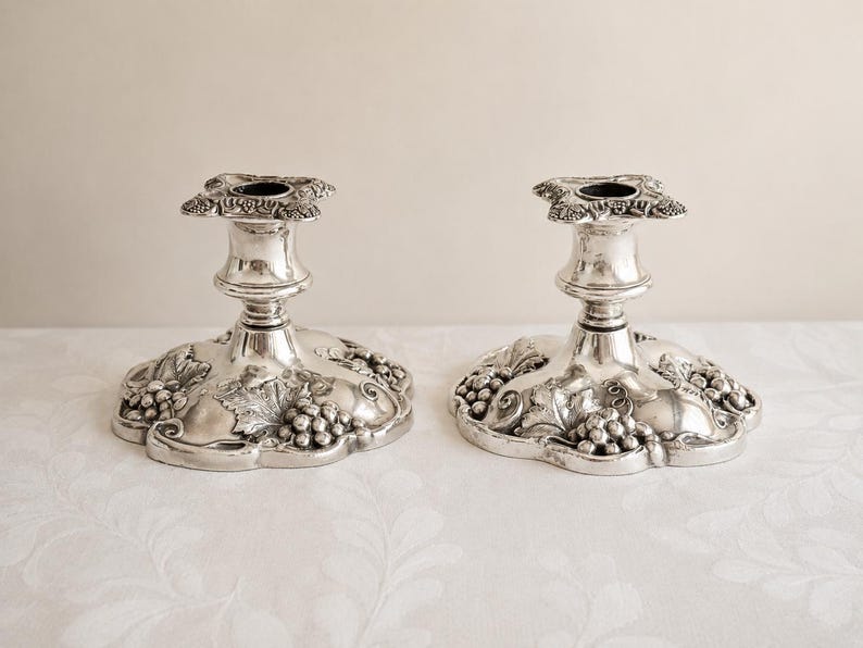 May include: A pair of silver-toned metal candle holders featuring a wide, scalloped base decorated with embossed grape clusters and vine leaf patterns. Each holder has a central stem and a square-shaped top rim with detailed floral accents.