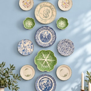 May include: A collection of twelve decorative plates arranged on a wall. The display includes blue and white transferware, floral patterned plates, and green cabbage-shaped plates. The plates vary in size and design, featuring intricate patterns.
