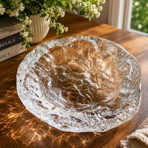 May include: A clear glass bowl with a textured, uneven surface that mimics the appearance of melting ice or flowing water. The bowl has a thick, irregular rim and a deep, rounded basin, reflecting light to create intricate patterns on the surface.