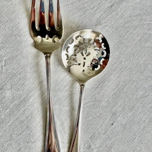 May include: Silver serving fork and spoon set. The fork has four tines with decorative cutouts, and the spoon has a round bowl with a floral design. Both have long, slender handles and are placed on a light-colored fabric.