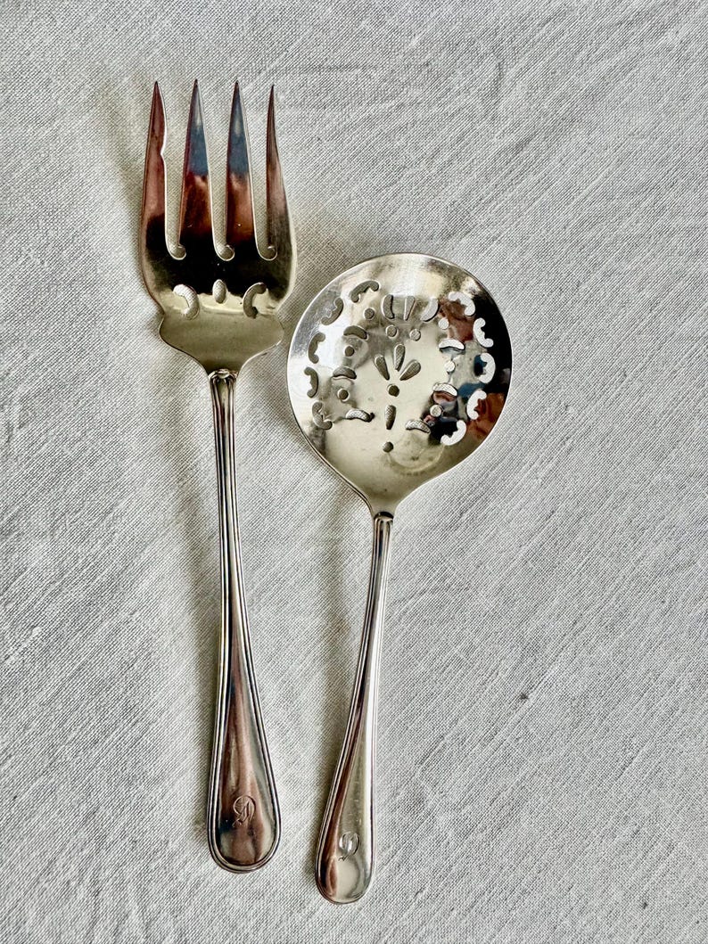 May include: Silver serving fork and spoon set. The fork has four tines with decorative cutouts, and the spoon has a round bowl with a floral design. Both have long, slender handles and are placed on a light-colored fabric.
