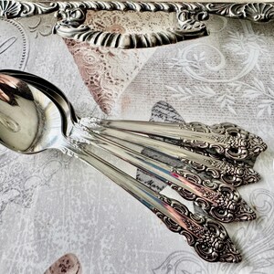May include: A collection of ornate silver spoons with detailed floral and scrollwork designs. The spoons are arranged on a patterned tablecloth, with a decorative silver tray in the background. The spoons are reflective and appear to be vintage.