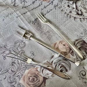 May include: Three silver serving forks on a floral fabric. The forks include a two-tine fork with a handle, a three-tine fork, and a standard fork. The fabric has a vintage design with a faded rose pattern.
