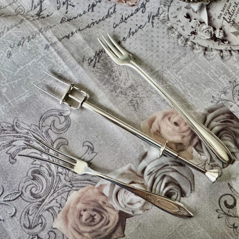 May include: Three silver serving forks on a floral patterned fabric. The forks vary in design, including a two-tine fork with a handle, a three-tine fork, and a standard fork. The fabric has a vintage aesthetic.