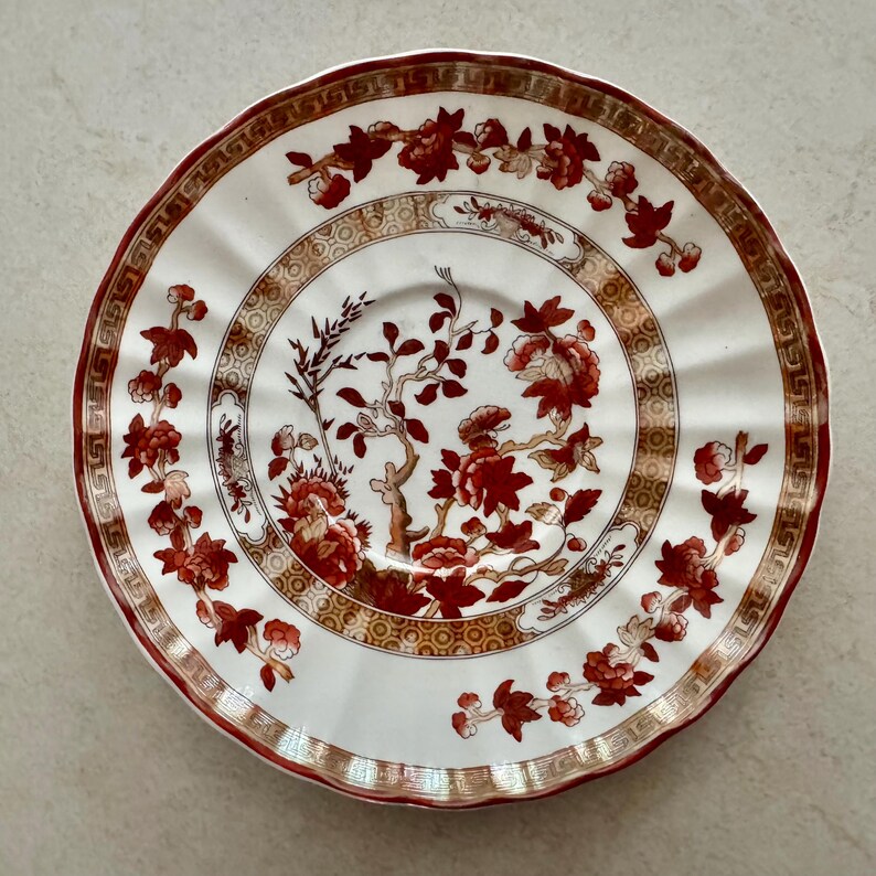 Pu&ograve; includere: Un piattino in ceramica rotondo con bordo smerlato, caratterizzato da una base bianca decorata con rami floreali rosso scuro e un motivo geometrico dorato sul bordo. Il centro mostra un dettaglio botanico con fiori rossi e steli.