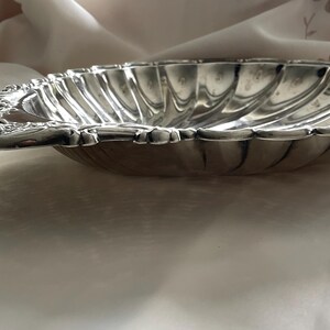 K&ouml;nnte beinhalten: Eine muschelf&ouml;rmige Schale aus silberfarbenem Metall mit gewelltem Rand und einem dekorativen Griff mit gepr&auml;gten Blumendetails. Das Innere der Schale weist ein geriffeltes Design auf, das von der Mitte zum geschwungenen Rand verl&auml;uft.
