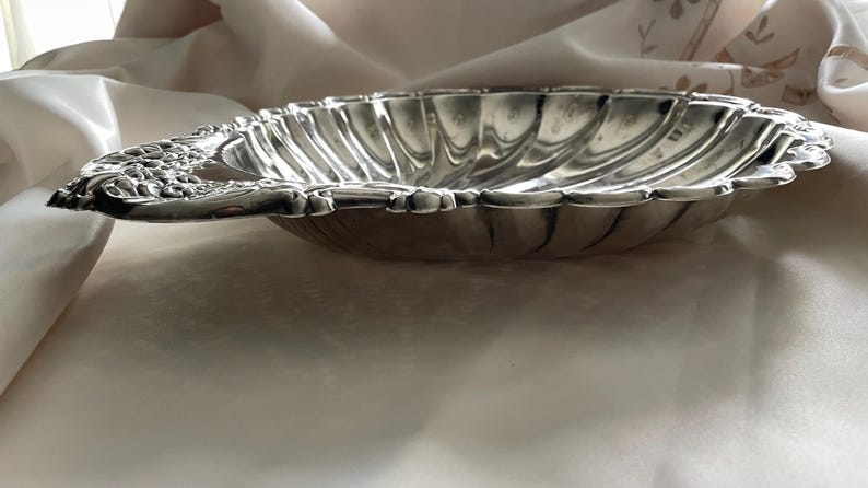 K&ouml;nnte beinhalten: Eine muschelf&ouml;rmige Schale aus silberfarbenem Metall mit gewelltem Rand und einem dekorativen Griff mit gepr&auml;gten Blumendetails. Das Innere der Schale weist ein geriffeltes Design auf, das von der Mitte zum geschwungenen Rand verl&auml;uft.