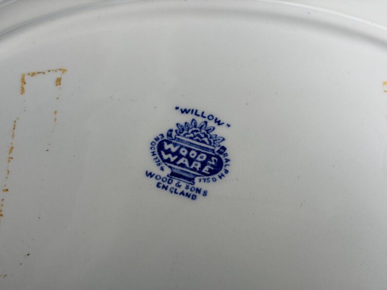 May include: A white ceramic plate base featuring a blue maker's mark in the center. The stamp displays the text "WILLOW," "WOOD & SONS," "ENGLAND," and "WOODS WARE" inside a decorative urn shape, with "ENOCH 1784" and "RALPH 1750" on the sides.