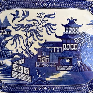 May include: A rectangular ceramic platter featuring a blue and white willow pattern. The design depicts a landscape with pagodas, a willow tree, two birds in flight, a bridge with figures, a small boat on water, and a decorative geometric border.