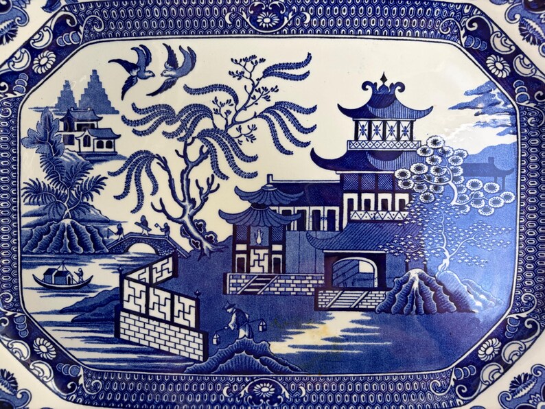 May include: A rectangular ceramic platter featuring a blue and white willow pattern. The design depicts a landscape with pagodas, a willow tree, two birds in flight, a bridge with figures, a small boat on water, and a decorative geometric border.
