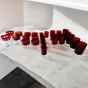 May include: A set of 28 red glass drinking glasses, including both stemmed and straight-sided styles. The glasses are arranged on a white surface.