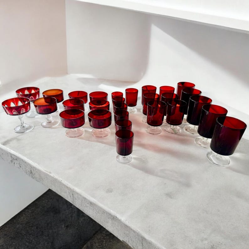 May include: A set of 28 red glass drinking glasses, including both stemmed and straight-sided styles. The glasses are arranged on a white surface.