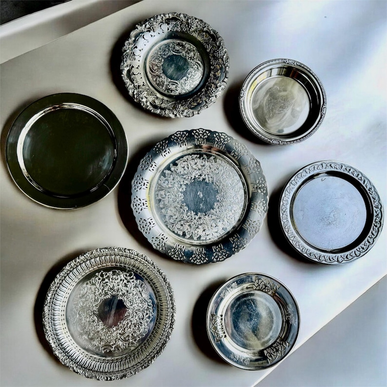 May include: A collection of seven silver-toned circular plates and trays of varying sizes. Some pieces feature intricate embossed floral and scrollwork patterns along the rims and centers, while others have smooth, polished surfaces with simple edges.