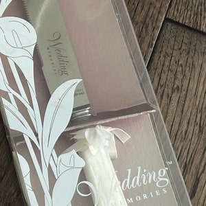 May include: A white wedding cake knife with a white satin ribbon bow and the words "Wedding Memories" on the blade. The knife is in a clear plastic box with a white floral design.