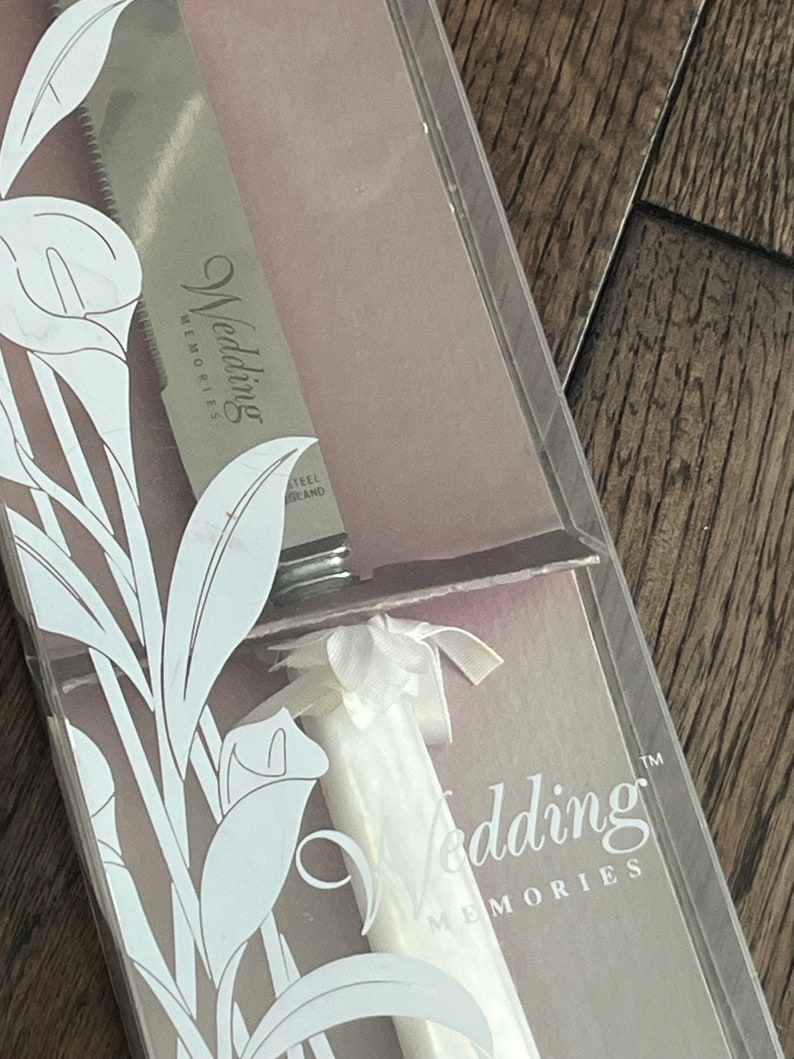 May include: A white wedding cake knife with a white satin ribbon bow and the words "Wedding Memories" on the blade. The knife is in a clear plastic box with a white floral design.