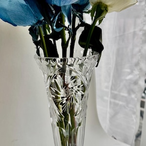 May include: A clear glass vase with a flared rim and a circular base, featuring intricate cut-glass starburst and vertical line patterns. The vase contains a bouquet of vibrant blue roses and white roses with green stems visible through the glass.