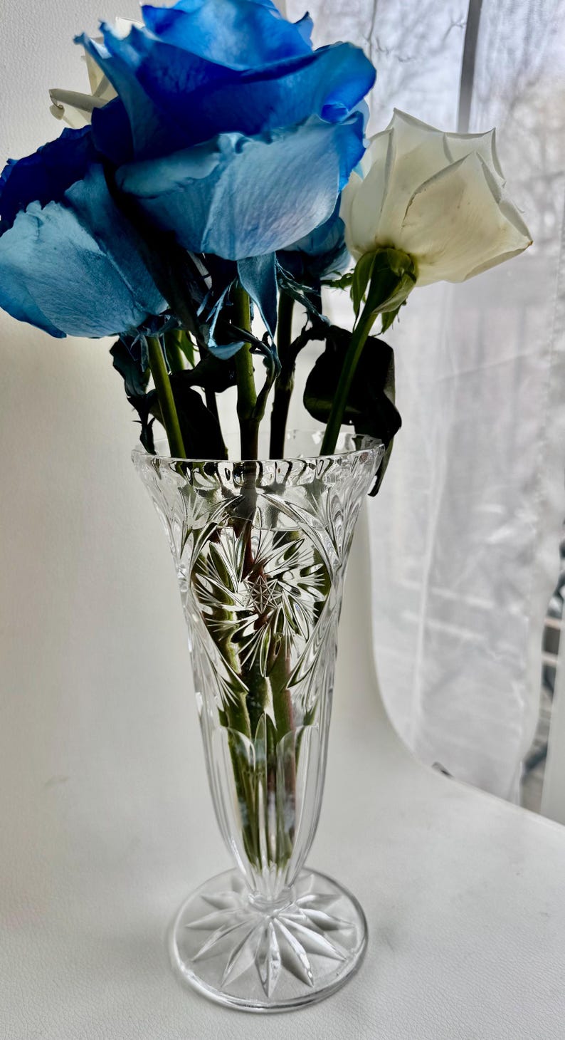 May include: A clear glass vase with a flared rim and a circular base, featuring intricate cut-glass starburst and vertical line patterns. The vase contains a bouquet of vibrant blue roses and white roses with green stems visible through the glass.