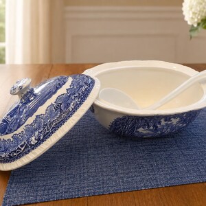 Op de afbeelding: Een witte keramieken soepterrine met een blauw transferware-patroon met een landschap. De set bevat een bijpassend deksel met een knopgreep en een witte keramieken soeplepel die in de kom met een geschulpte rand ligt.