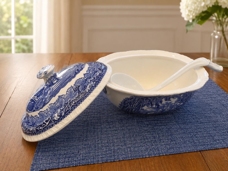 Op de afbeelding: Een witte keramieken soepterrine met een blauw transferware-patroon met een landschap. De set bevat een bijpassend deksel met een knopgreep en een witte keramieken soeplepel die in de kom met een geschulpte rand ligt.
