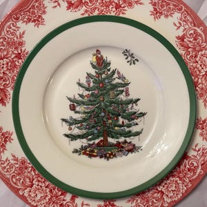以下が含まれることがあります： 緑色の縁取りのある白いセラミック製のプレートには、赤、緑、金色のオーナメントがついたクリスマスツリーのデザインが施されています。プレートは、花柄の大きな赤と白のプレートの上に重ねられています。