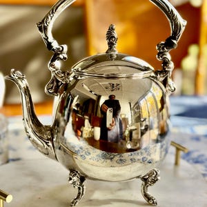 May include: A polished silver-toned teapot featuring an ornate, curved handle, a decorative spout, and a lid with a finial. The rounded body stands on three detailed, claw-style feet, with a reflective surface showing a distorted room interior.