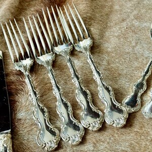 May include: A set of silver flatware, including forks and a knife, with ornate handles. The flatware is laid out on a brown fur surface. The forks have the number "101" stamped on the back of the handle.
