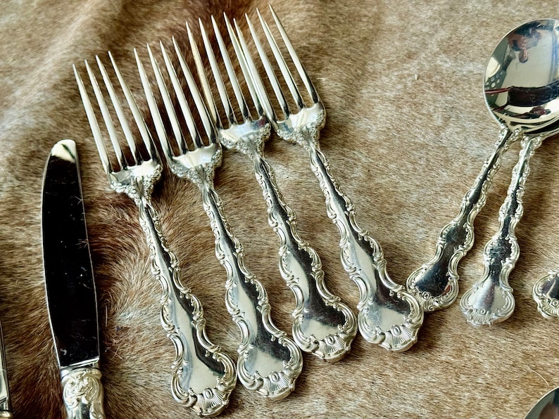 May include: A set of silver flatware, including forks and a knife, with ornate handles. The flatware is laid out on a brown fur surface. The forks have the number "101" stamped on the back of the handle.