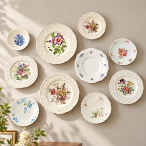 May include: A collection of nine ceramic plates arranged on a wall, featuring various floral patterns, scalloped edges, and embossed textures. The plates display colorful botanical illustrations including roses, tulips, and violets on white backgrounds.