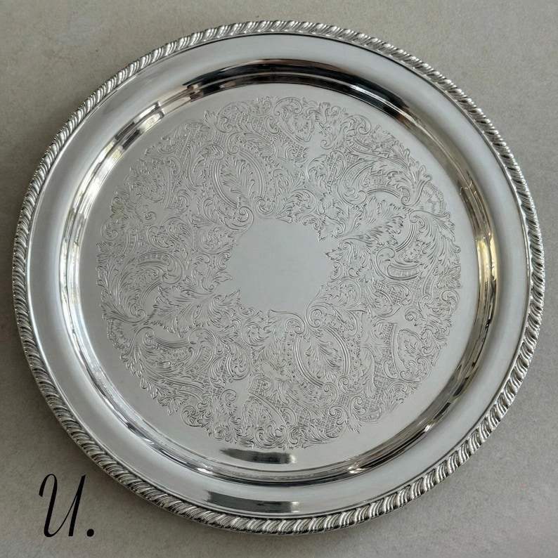 May include: A round silver-toned serving tray featuring a polished finish and a decorative rope-style border. The center of the tray is adorned with an intricate, engraved floral and scrollwork pattern surrounding a plain circular area.