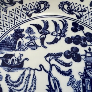 K&ouml;nnte beinhalten: Wei&szlig;e Keramikplatte mit blauem Willow-Muster. Die Platte zeigt eine detaillierte Landschaftsszene mit Pagoden, V&ouml;geln und einem Boot. Der Rand hat ein geometrisches Muster und eine Scroll-Muster.