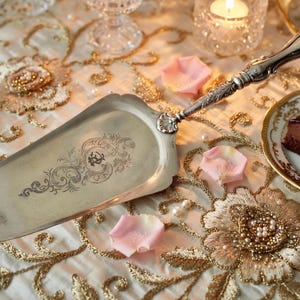 K&ouml;nnte beinhalten: Ein silberner Tortenheber mit einem verzierten Griff und dekorativer &Auml;tzung. Der Heber liegt auf einer gold-cremefarbenen, bestickten Tischdecke neben einem St&uuml;ck Schokoladenkuchen mit einer Himbeere.