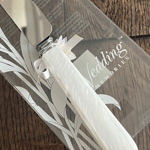 May include: A white cake knife with a silver blade and a white handle with a floral design. The knife is in a clear plastic box with a white background and the words "Wedding Memories" printed on it.