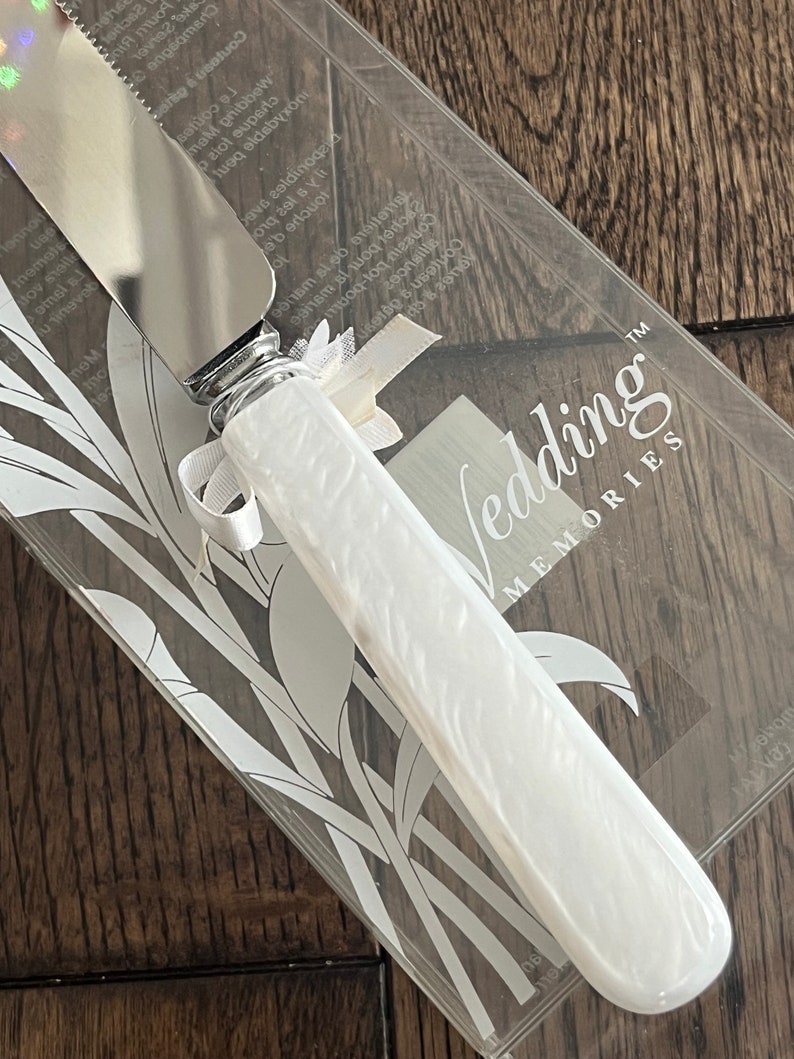 May include: A white cake knife with a silver blade and a white handle with a floral design. The knife is in a clear plastic box with a white background and the words "Wedding Memories" printed on it.