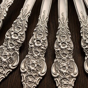 May include: A set of silver-colored metal spoons featuring intricate embossed floral patterns on the handles. The design includes detailed flowers, leaves, and scrolling elements that extend from the base up the stem of each individual utensil.