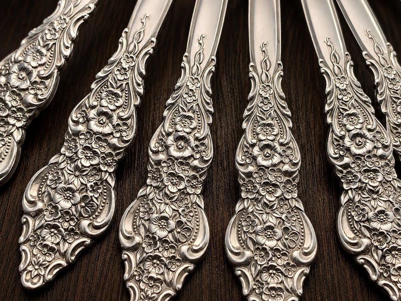 May include: A set of silver-colored metal spoons featuring intricate embossed floral patterns on the handles. The design includes detailed flowers, leaves, and scrolling elements that extend from the base up the stem of each individual utensil.