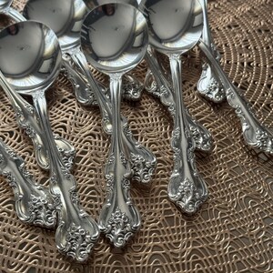 May include: A collection of ornate silver soup spoons. The spoons have a reflective, polished surface and decorative floral patterns on the handles. The spoons are arranged on a textured, gold-colored placemat.